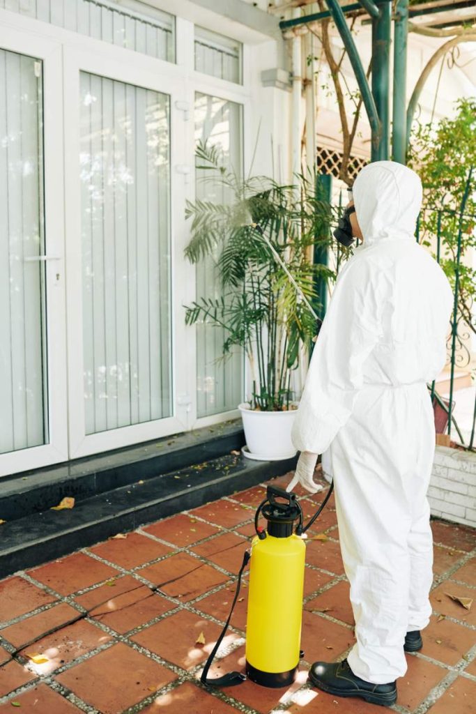technician in hazmat suit spraying chemicals on walls and windows of house to prevent virus spread