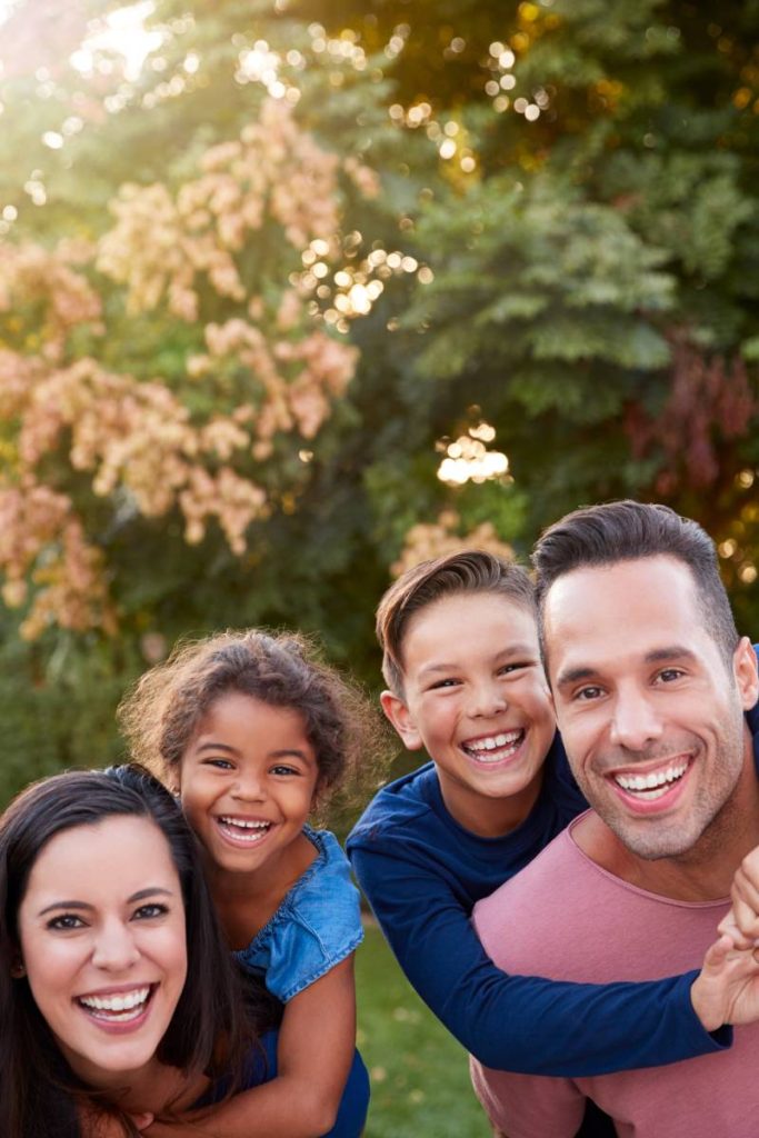 Portrait Of Smiling Hispanic Family With Parents Giving Children Piggyback Rides In Garden At Home