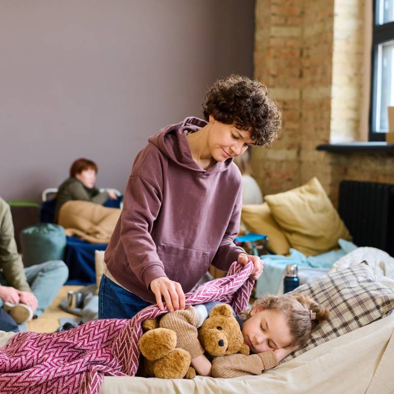Young brunette woman covering her sleeping son with teddybear with warm blanket against other temporarily homeless people
